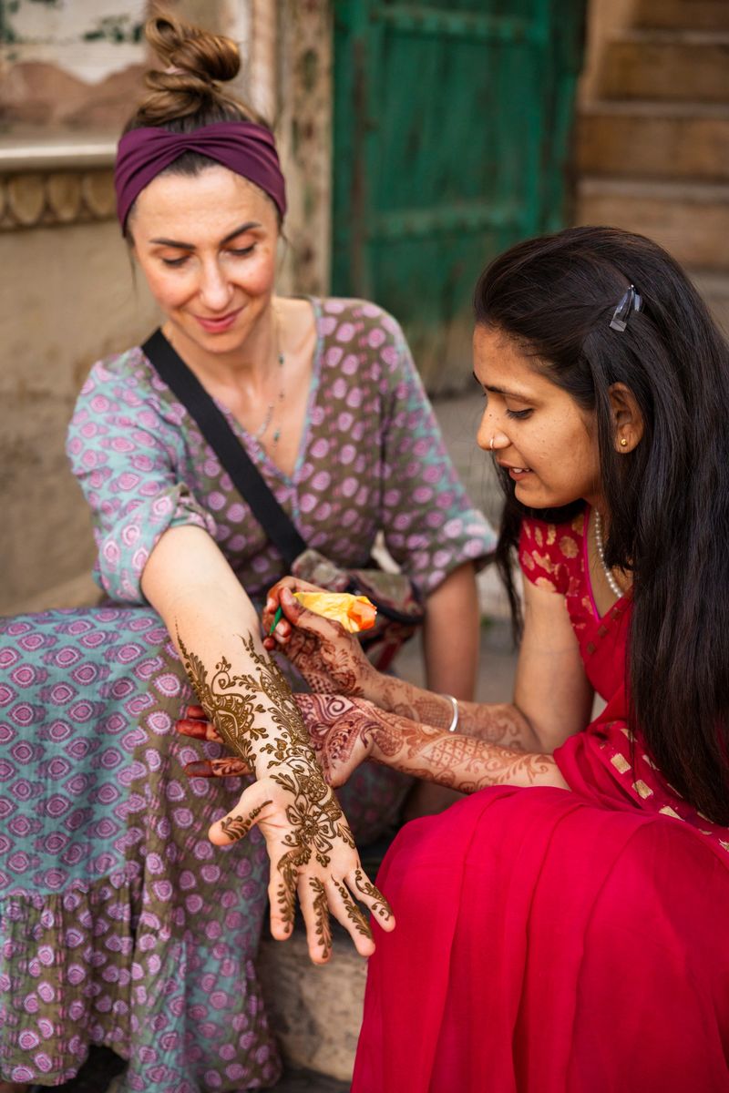 A local henna artist in a red sari carefully applies intricate mehndi designs to the outstretched forearm and hand of a smiling visitor wearing a patterned dress and headband. They sit outdoors by a rustic doorway and stone steps as the artist concentrates on creating floral and paisley motifs, the detailed brown paste contrasting with the visitor’s skin and the colorful clothing.