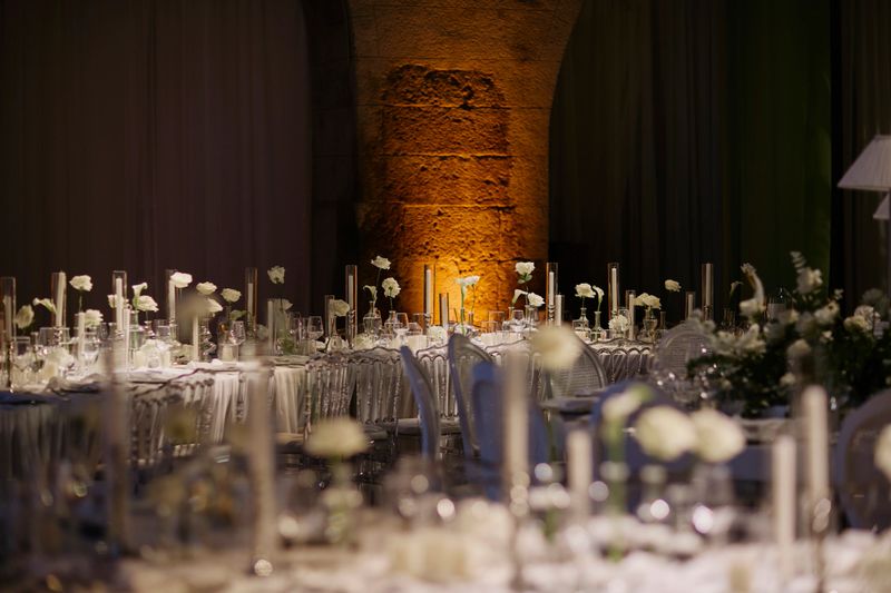 Elegant wedding reception tables arranged inside a historic stone hall, featuring white floral details, tall candles, refined glassware, and transparent chairs. Warm architectural lighting highlights the textured stone columns, creating a romantic and sophisticated atmosphere. Ideal for concepts of luxury weddings, destination venues, fine dining events, historic interiors, and upscale celebration design. stock photo