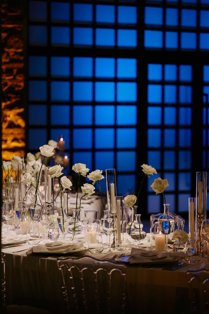 Elegant wedding reception tables decorated with white floral arrangements and glowing candles inside a modern venue. Blue backlit architectural windows create a dramatic contrast with the warm candlelight, enhancing the sophisticated evening atmosphere. Ideal for concepts of luxury weddings, contemporary event design, fine dining, upscale celebrations, and romantic interior styling. stock photo