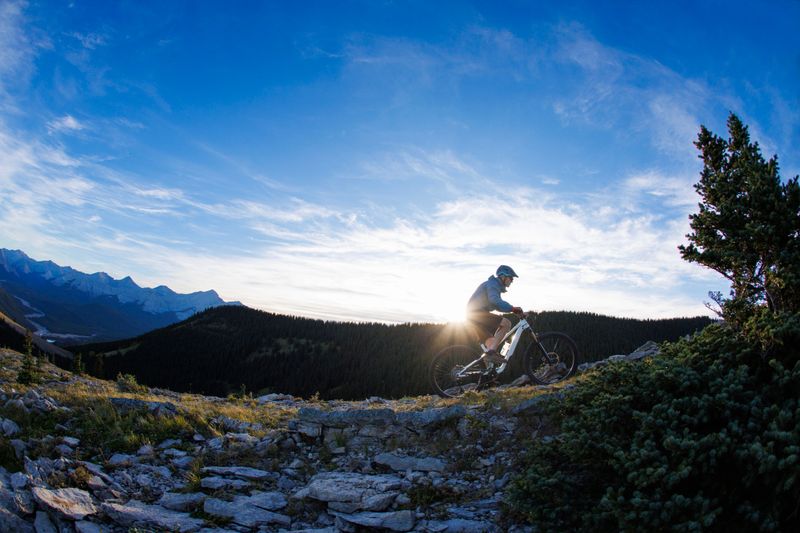 A man rides his full-suspension, electric-powered mountain bike up a trail in the Rocky Mountains of Alberta, Canada, at sunset. He wears a warm jacket, bicycle shorts, sunglasses, and a bicycle helmet.