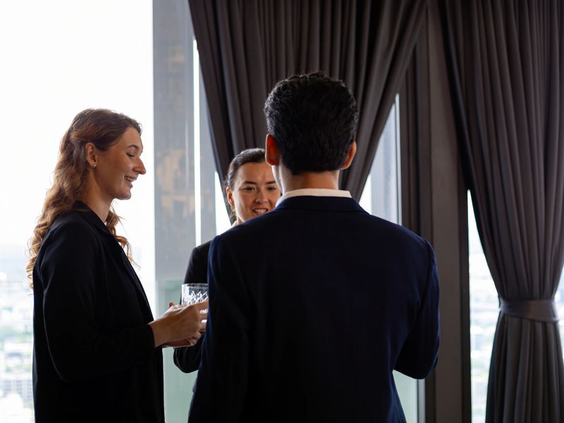 Three business professionals networking and talking together near window in modern office holding drinks showing communication confidence teamwork and corporate culture