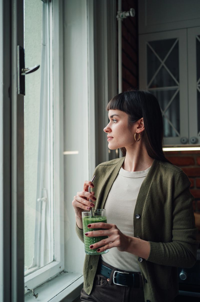 Young woman standing by window holding a healthy green smoothie, looking away while enjoying a healthy lifestyle moment