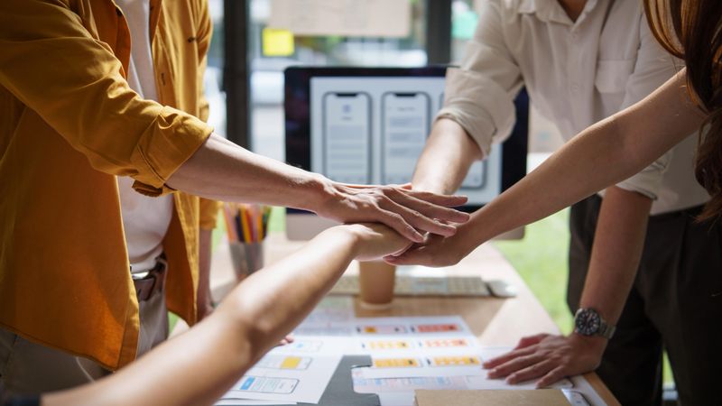 Smiling team members stacking hands over a design table, celebrating teamwork and shared goals in a UX UI workspace.