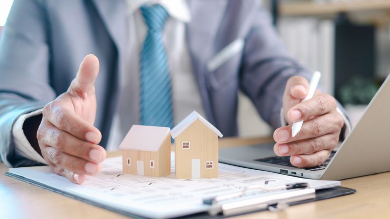 Businessman protecting two house models on documents while working on laptop. Concept of real estate investment, property portfolio, home insurance, and financial planning.