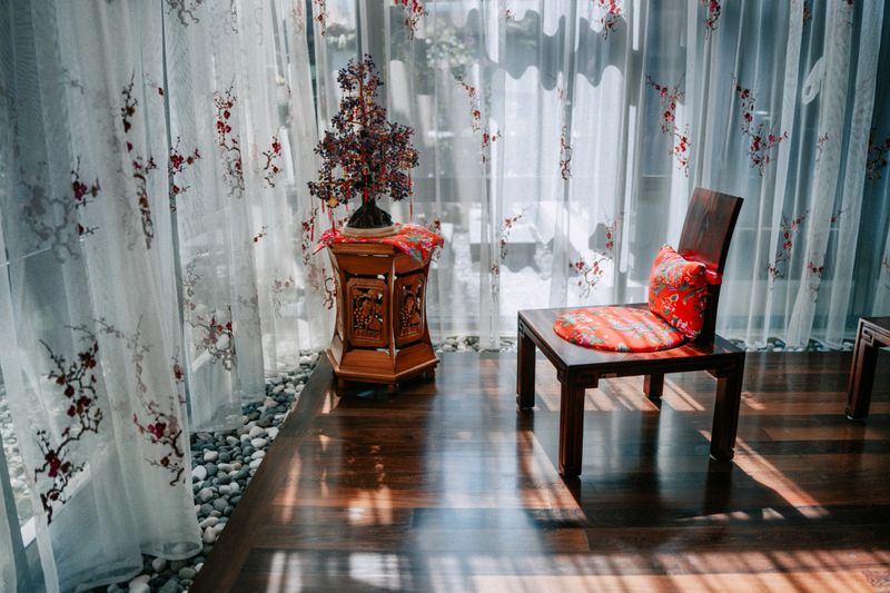 Sunlight streams through sheer curtains onto a wooden floor beside a traditional chair and side table, creating a calm home interior scene with copy space,  Kuala Lumpur