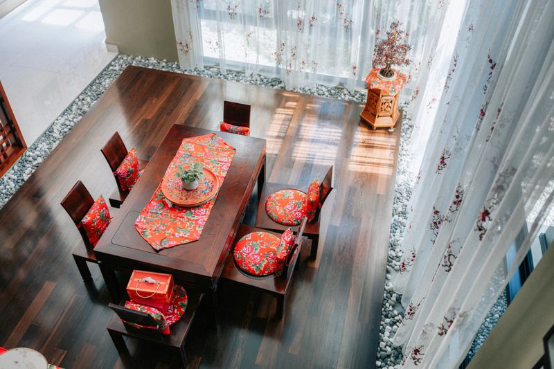 A top-down view of a wooden dining table arranged for celebration with red embroidered textiles in a bright home interior with sunlight, sheer curtains, and a crystal tree decoration,