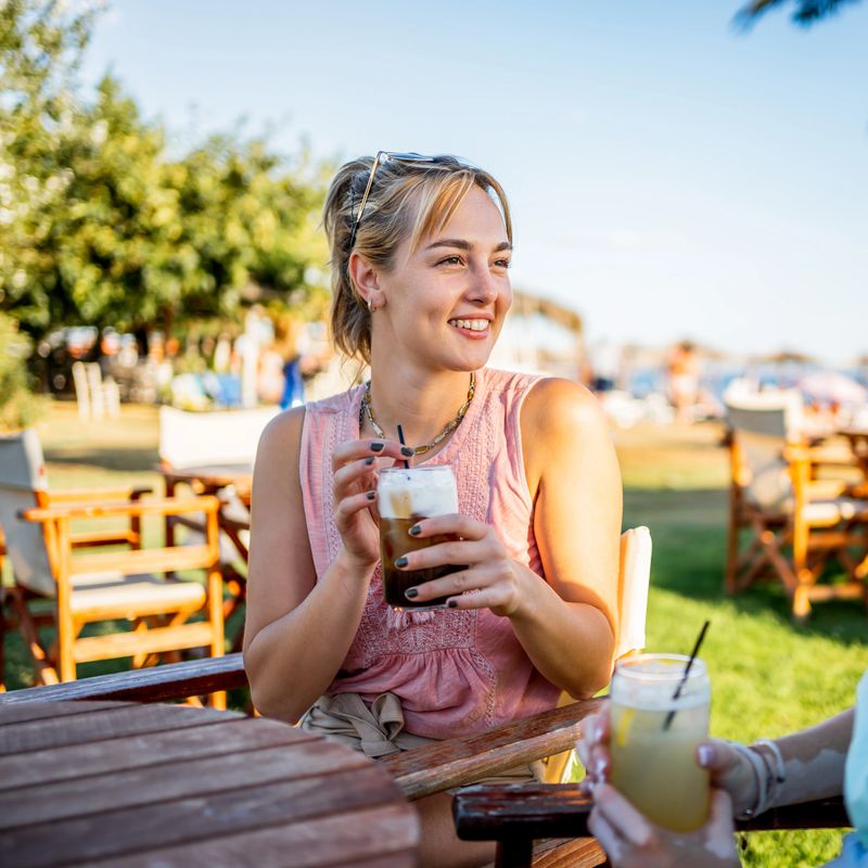 Young woman smiling, holding an iced coffee, and relaxing with a friend at an outdoor cafe near the beach