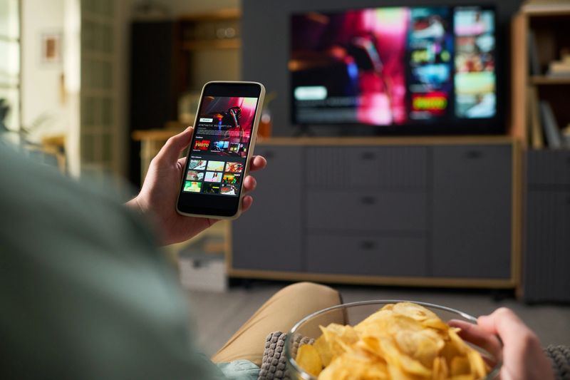 Unknown man holding smartphone while browsing streaming service app, sitting on sofa with bowl of potato chips, watching television screen in modern living room