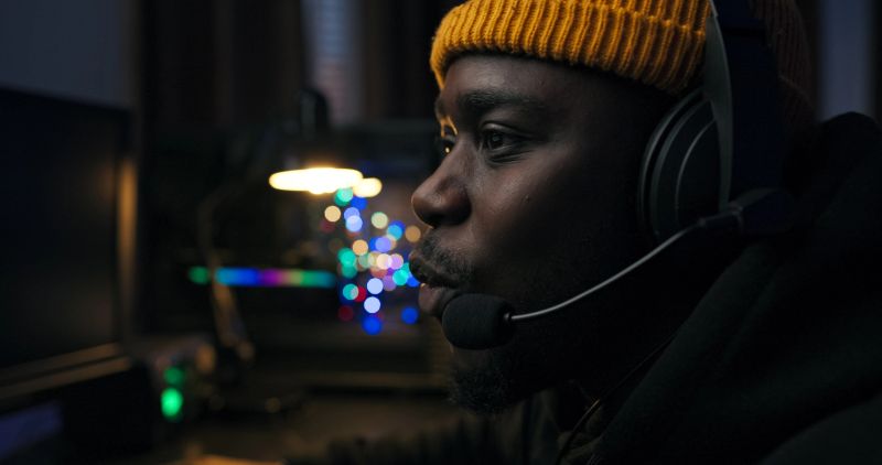 Focused male player relives gaming rounds in dark room. Colorful LED lights surround him, tapping keyboard quickly, emotional reactions flash across his face.