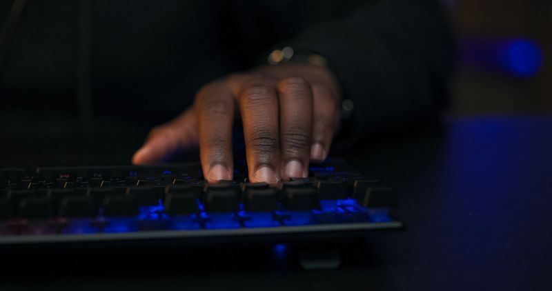 Close-up of a male gamer pressing colorful backlit keyboard keys, immersed in gameplay. LED lights reflect on his hands as he focuses on a fast-paced computer game.
