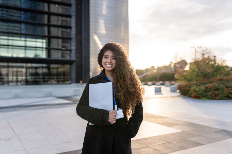 Young professional woman smiling, holding a laptop and credentials outside a modern office building, ready for business and success