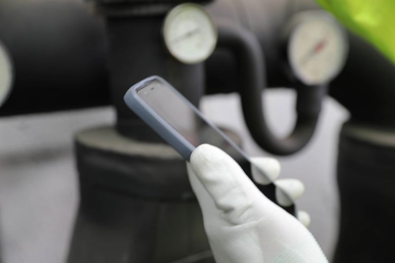 Industrial Technician Wearing Protective Clothing And Safety Equipment Using A Smartphone To Check Pressure Gauge And Monitor Heating Or Cooling System In A Technical Facility, Performing Digital Inspection And Maintenance Control.
