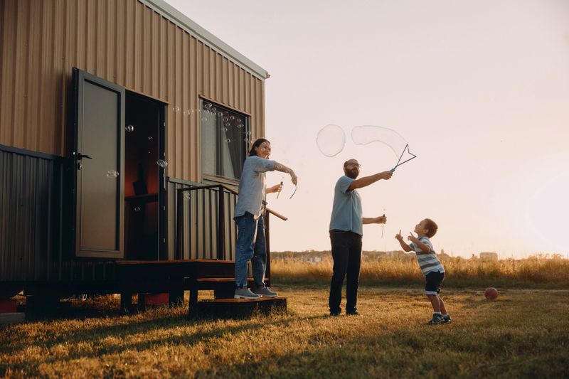 A family of three enjoys a playful evening outside their modern tiny house, blowing bubbles as the sun sets. Adult parents and a minor child share joyful moments in a warm outdoor setting.