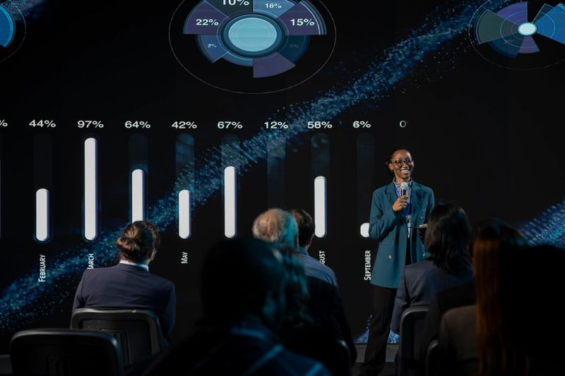 Stage presentation at international business event with diverse participants, networking pitch in conference hall. Black female speaker addressing audience for startup development.