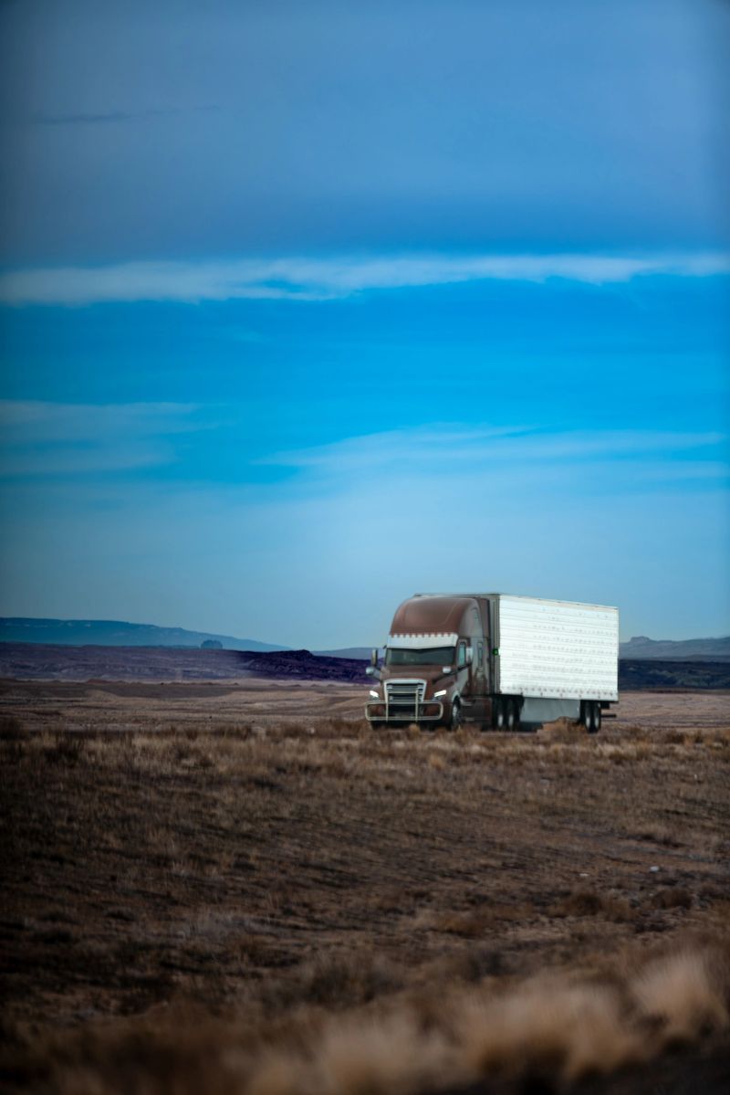 A large commercial semi-truck with a white trailer travels across a barren, brown landscape under a vibrant blue sky. The truck is positioned in an open, arid terrain with mountain ranges visible in the background, creating a sense of isolation and long-distance transportation.