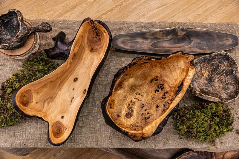A collection of uniquely shaped, handcrafted wooden bowls and plates displayed on burlap with moss accents. The bowls exhibit natural grain patterns and organic forms