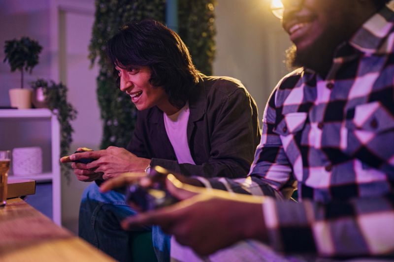 Diverse young adult male friends laughing and holding game controllers while enjoying a competitive video game match in a living room at night
