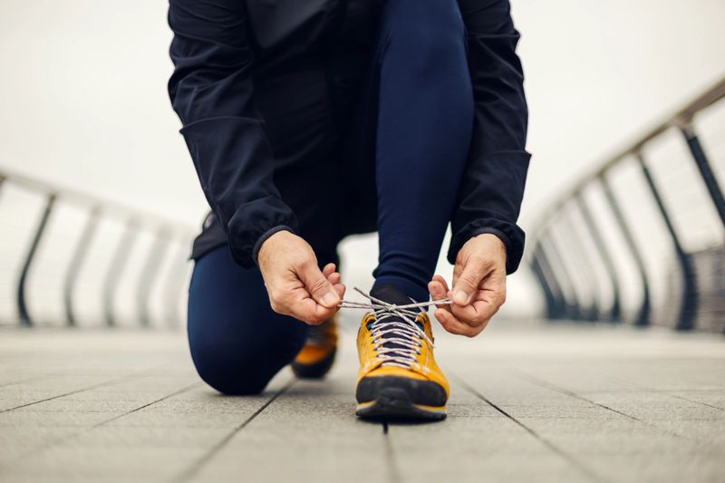 Mid-adult runner bending on an urban bridge path tying bright yellow running shoe, close-up low-angle shot conveying preparation, determination and outdoor fitness routine
