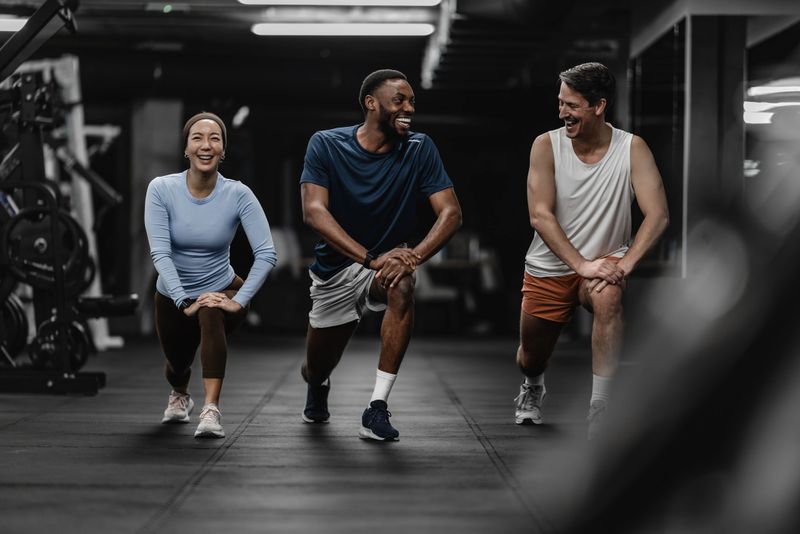 A diverse trio works out in a modern gym, sharing smiles and energy during lunges. The scene highlights fitness, friendship, and motivation in a dynamic group workout.