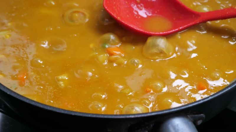 A bubbling yellow curry sauce simmers in a large black pan containing diced potatoes, carrots, corn, and protein bits to create a hearty and aromatic traditional meal in a professional kitchen.