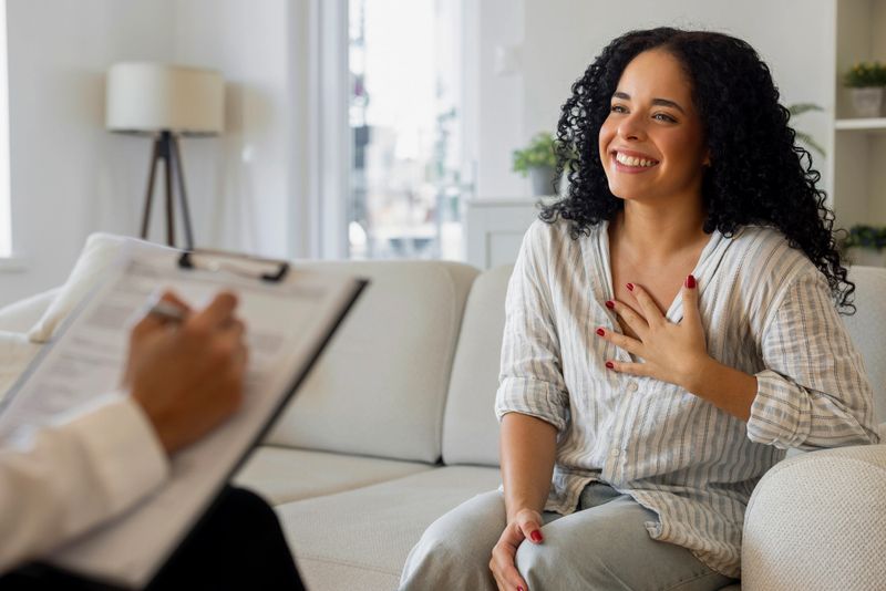 Portrait of a happy and relieved young woman with curly hair sitting on a sofa during a psychotherapy session. She is smiling broadly and touching her chest with gratitude while talking to a counselor or psychologist who is taking notes in the blurred foreground. The scene represents successful mental health treatment, recovery, and positive emotional breakthrough.