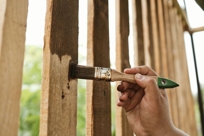 A person is carefully staining vertical wooden planks, protecting them from the elements with a rich, dark finish