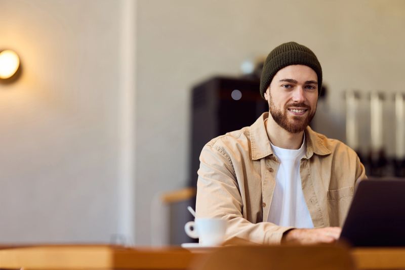 A young man with a beanie wearing a beige shirt and white tee sits at a cafe table, typing on a laptop beside a coffee cup, conveying focused work, remote productivity, and relaxed modern lifestyle.