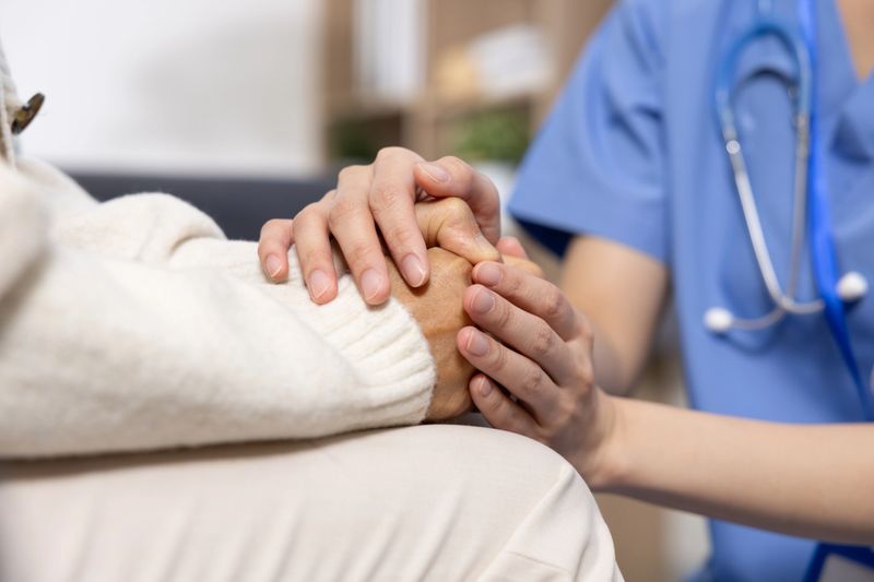 Asian Compassionate healthcare worker holding elderly patient hand, showing care and support in medical setting with stethoscope visible on uniform
