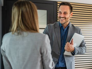 Two professionals in a friendly office conversation, one holding a laptop.