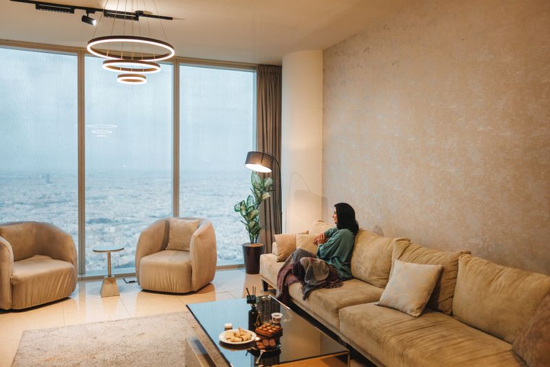 Mid adult Arabic female relaxing on a comfortable sofa in a luxury living room featuring a panoramic view of Riyadh cityscape. She appears calm and content, surrounded by plush furniture and elegant decor.