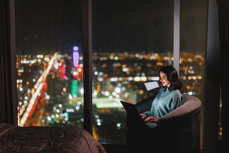 Mid adult Arabic businesswoman seated in a luxury apartment in Riyadh, using a laptop while overlooking the cityscape at night. She is focused and engaged, wearing a button-up blouse. The room provides a stunning view through large windows, highlighting the vibrant city lights.