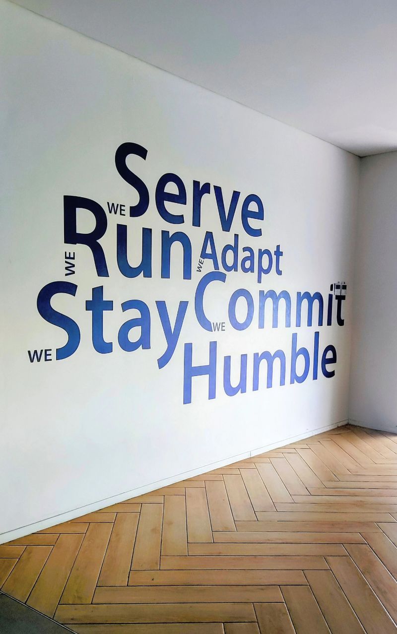 Interior shot of a modern workspace featuring large blue typography on a white wall. The words display corporate values such as "We Serve, Run, Adapt, Stay, Commit, Humble." The floor is made of wood with a herringbone pattern. Perfect for business culture and teamwork themes.