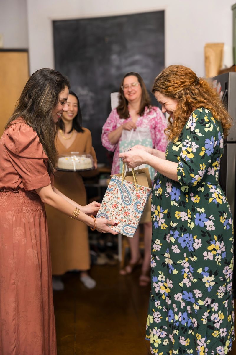 A group of women gather for a baby shower celebration in a modern home interior. Two women in floral dresses exchange a patterned gift bag while friends look on with smiles, creating a warm atmosphere of friendship and anticipation. The scene captures the joy and tradition of celebrating new life with loved ones during this special milestone event.