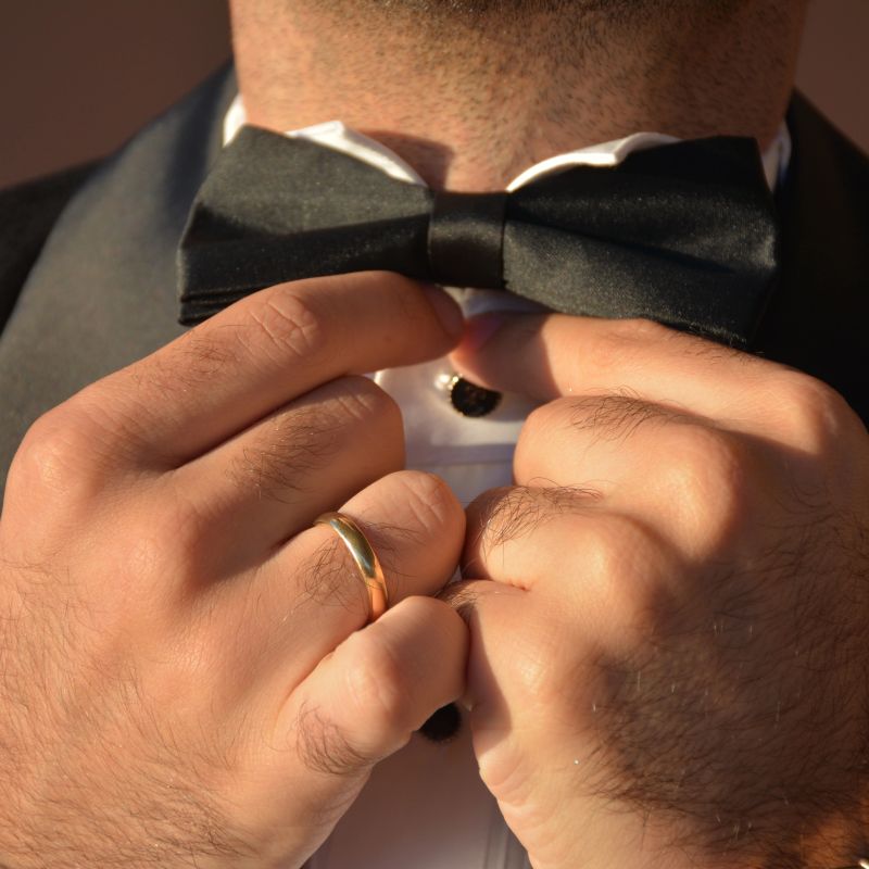 Groom Adjusting Black Bow Tie Close Up Preparation For Wedding Ceremony. Wearing a black tuxedo suit and a white shirt with black buttons. Detail of hands with a gold wedding ring during preparation.