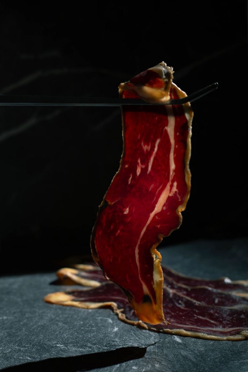 A slice of Spanish cured beef cecina delicately held with metal tweezers against a dark background, illuminated to reveal its glossy surface and rich translucent texture. High-end artisanal charcuterie presented in a moody, elegant atmosphere, emphasizing exclusivity, craftsmanship and refined Mediterranean gastronomy.