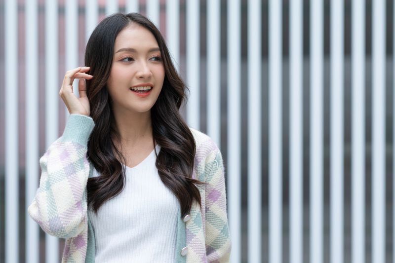 Asian Young woman smiling with long wavy hair wearing pastel checkered cardigan and white top standing in front of striped background, expressing happiness and confidence