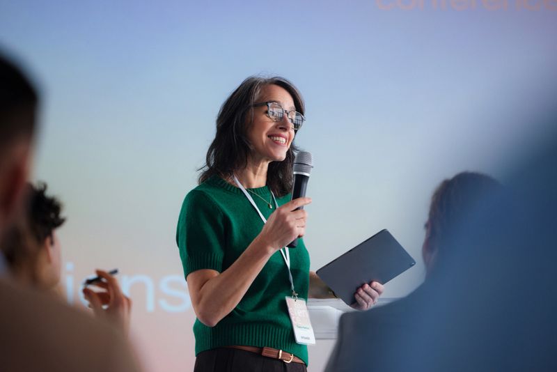 A confident female speaker in a green sweater delivers a talk at a conference, holding a microphone and tablet while engaging a focused audience in a bright, professional setting.