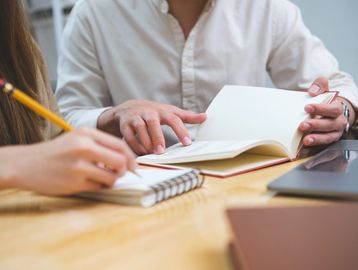 Two people studying together, one writing notes and the other reading a book.