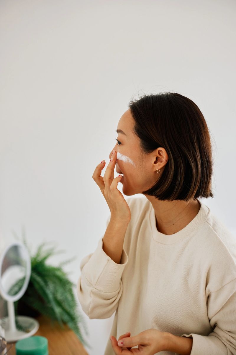 A beautiful Asian woman performs a morning skincare routine at home, applying cream to her face with calm focus. Soft natural light, cozy living space, and a relaxed, self-care mood.