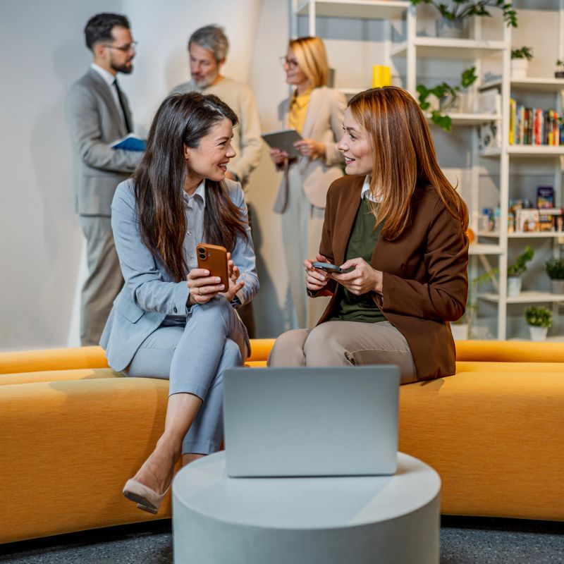 Two businesswomen sitting, talking, and smiling while using mobile phones in a vibrant office lounge area