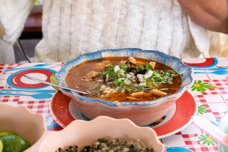 A person is seated at a table, enjoying a bowl of traditional soup filled with various ingredients. The dining setting includes bright colors and cheerful decorations.