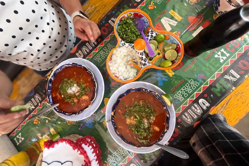 Two bowls of traditional Comida Mexicana are on a colorful table. People are enjoying the food with various toppings like chopped onions and lime. Drinks are nearby.