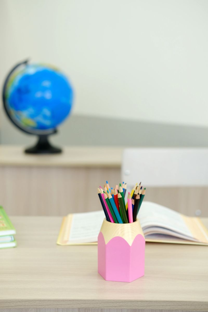 Vibrant colored pencils arranged in a pink holder on a wooden school desk, with an open book and globe nearby, enhancing the learning environment