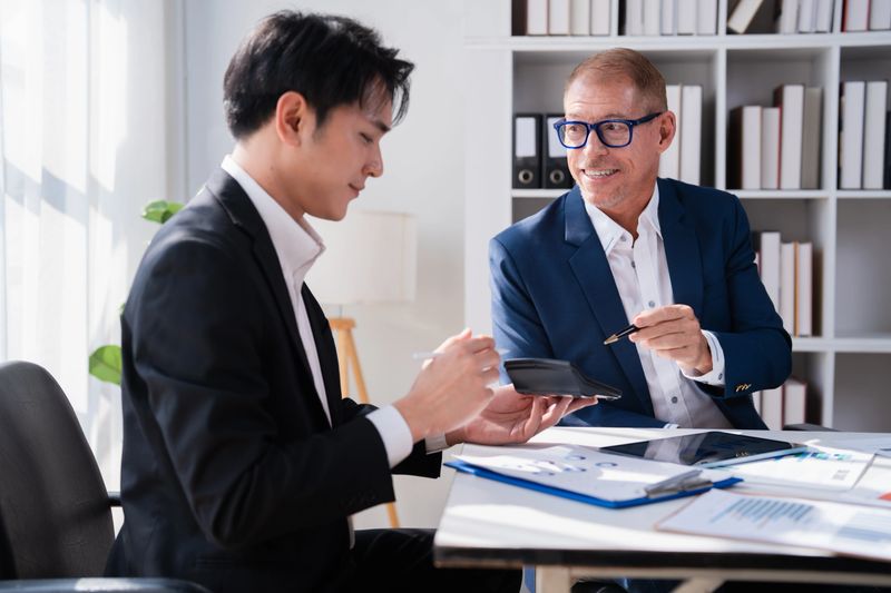 Two businessmen are collaborating on a financial plan, reviewing documents and using a calculator while discussing investment strategies in a bright, contemporary office environment