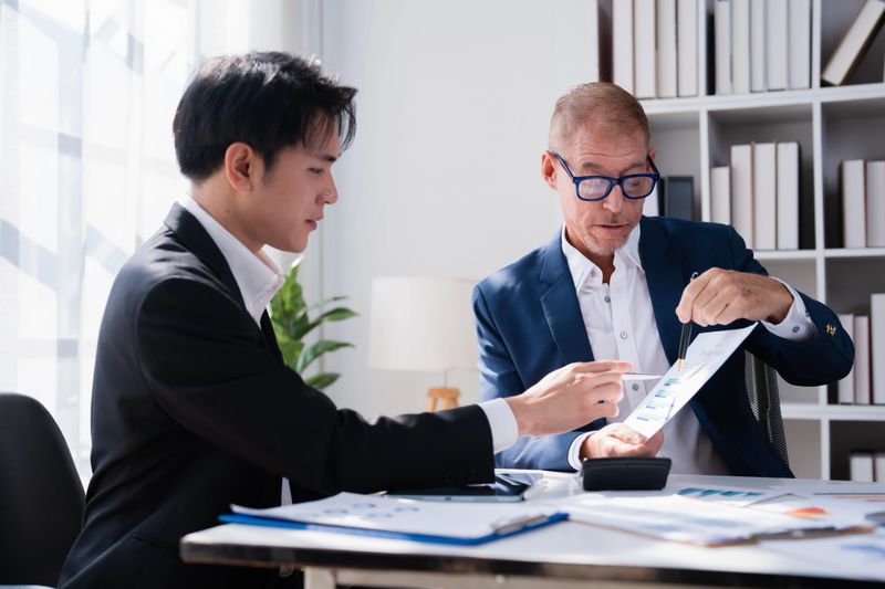 Two businessmen collaborating at an office desk, analyzing financial graphs and charts on documents to understand market trends and develop new strategies