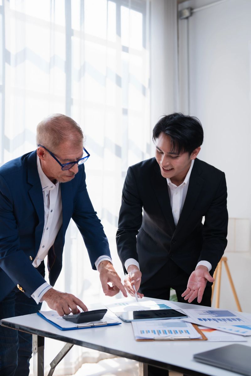 Two businessmen are collaborating on a financial strategy, analyzing charts and documents on a table in a bright office, discussing data and calculating figures during a meeting