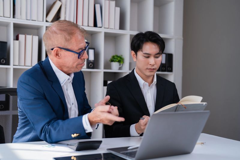 Two diverse businessmen collaborating on a project, with one experienced professional providing guidance and advice to a younger colleague during a business meeting in their modern office