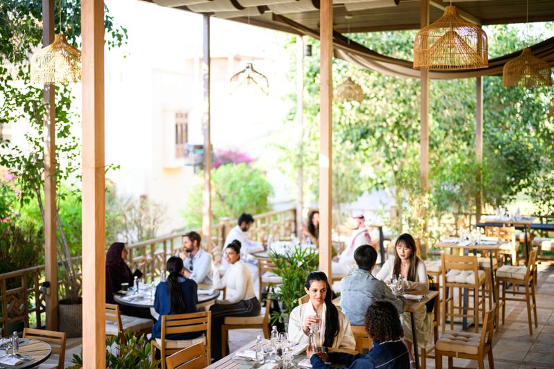 A diverse group of people sitting at tables, enjoying drinks and conversation during a bright daytime meal on a spacious outdoor restaurant patio. Customers relaxing under a shaded wooden structure surrounded by lush green foliage and natural light. The setting provides a comfortable, upscale environment for socializing.