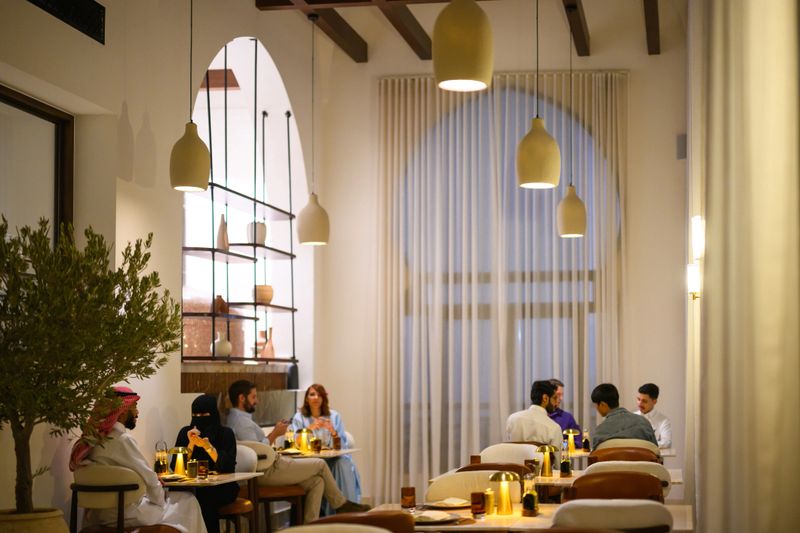 Diverse group of people sitting at round tables, conversing and enjoying dinner service in a luxurious Middle Eastern hotel restaurant. Soft, warm ambient light illuminating the modern minimalist decor and natural materials, creating a comfortable and exclusive evening dining experience. Arched windows provide architectural interest and contrast.