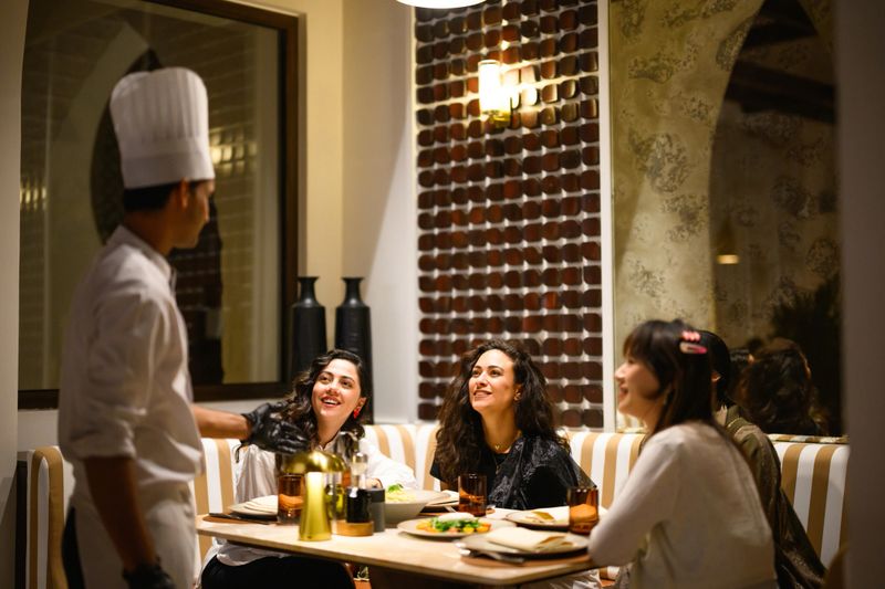 An Arab chef in traditional attire personally serving a diverse group of Arab women seated at a table in an elegant, dimly-lit restaurant setting. The women are engaged in conversation while enjoying their meal, with decorative lighting and architectural details creating a warm, inviting atmosphere.